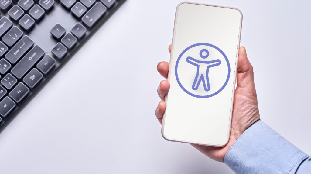 Un teclado encima de una mesa blanca y encima de ello una mano sosteniendo un celular y dentro de el el simbolo de accesibilidad.